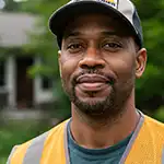 A headshot of Marcus Thorne, in a baseball cap and safety vest, smiling at the camera outdoors.